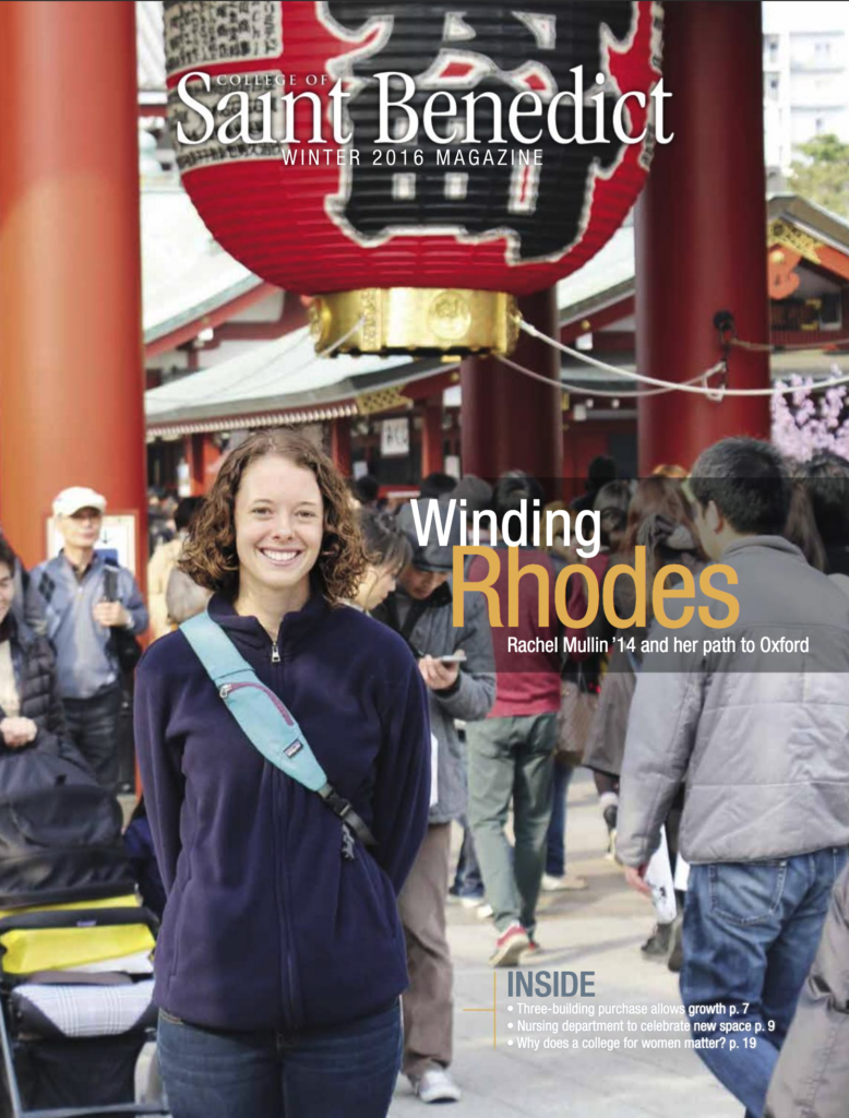 A woman smiling at the camera stands in a crowded outdoor marketplace, holding a drink. She is wearing a blue jacket and a shoulder bag. In the background, there are traditional structures and various people walking.